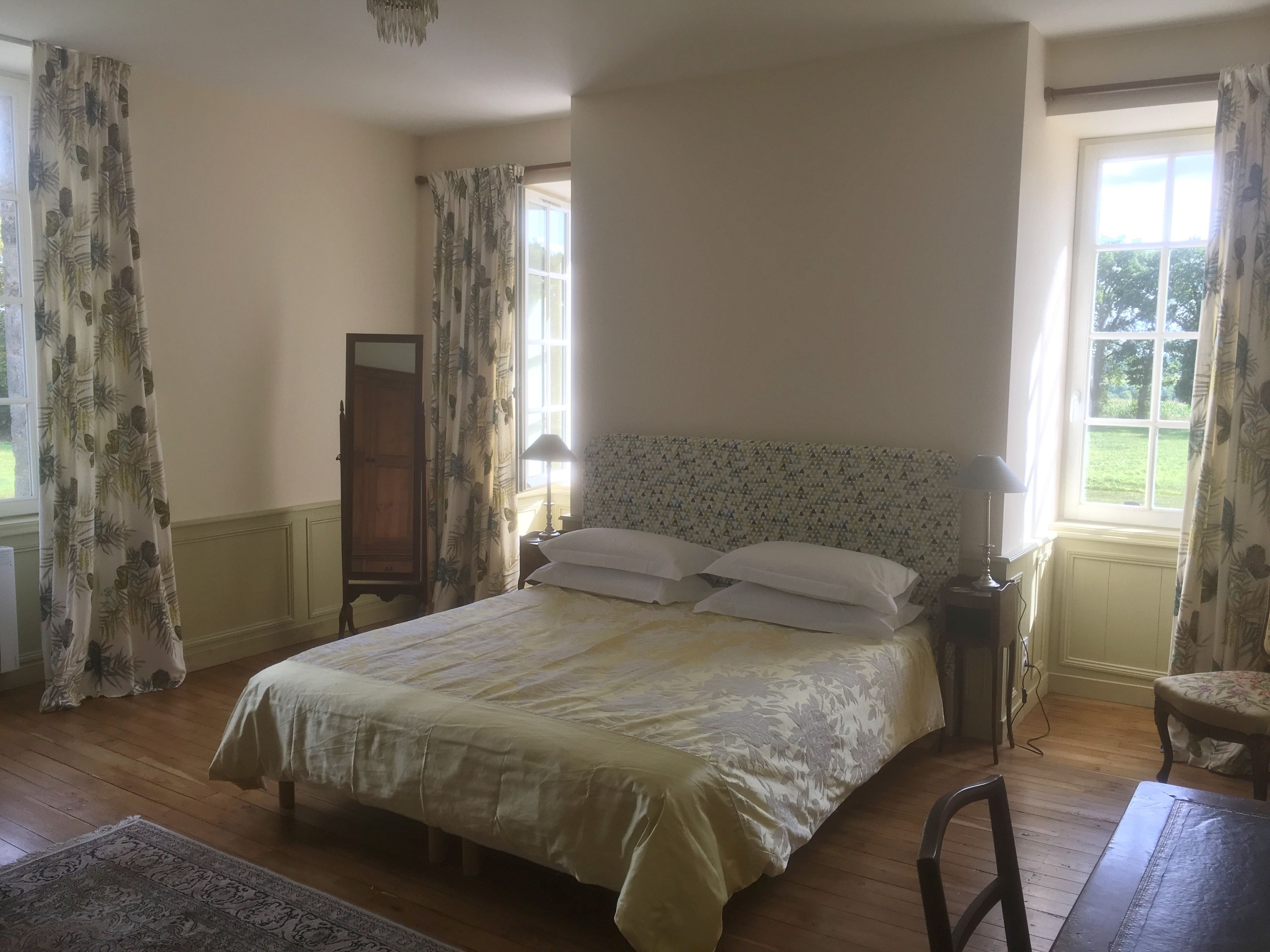 A bedroom with a large bed, a nightstand with a lamp, a wooden wardrobe, and a window with curtains.