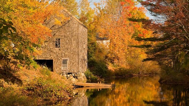 A rustic stone building with a dock extending into a calm river, surrounded by autumn foliage.