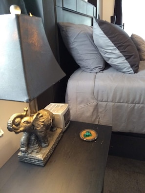 A bed with grey bedding and a black headboard, a nightstand with a lamp, a small elephant figurine, and a microwave. - The Cottages of Lynchburg (Lynchburg)