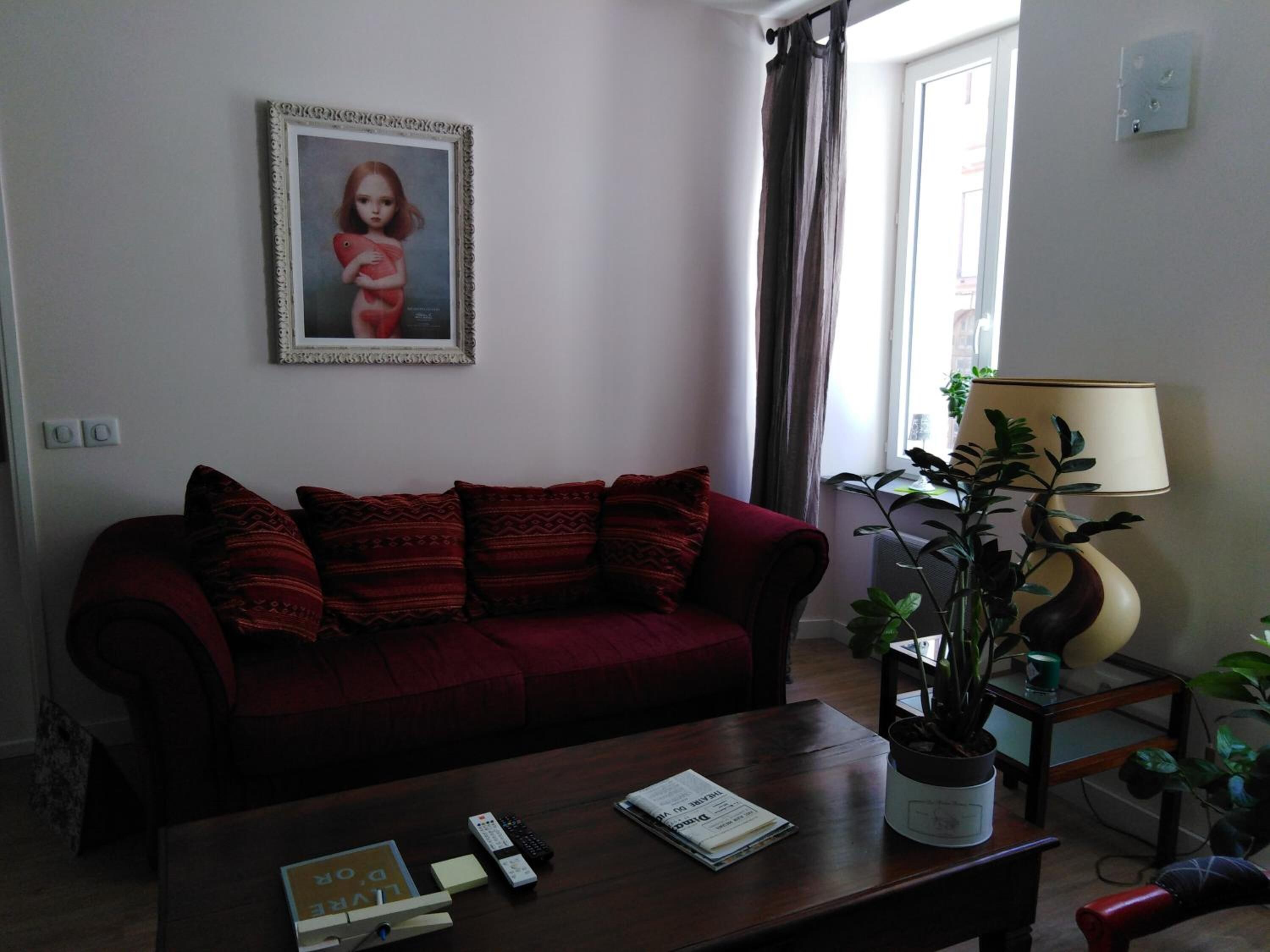 Un salon avec un canapé rouge, une table basse sur laquelle se trouvent des télécommandes et un livre, une lampe et un tableau encadré au mur.