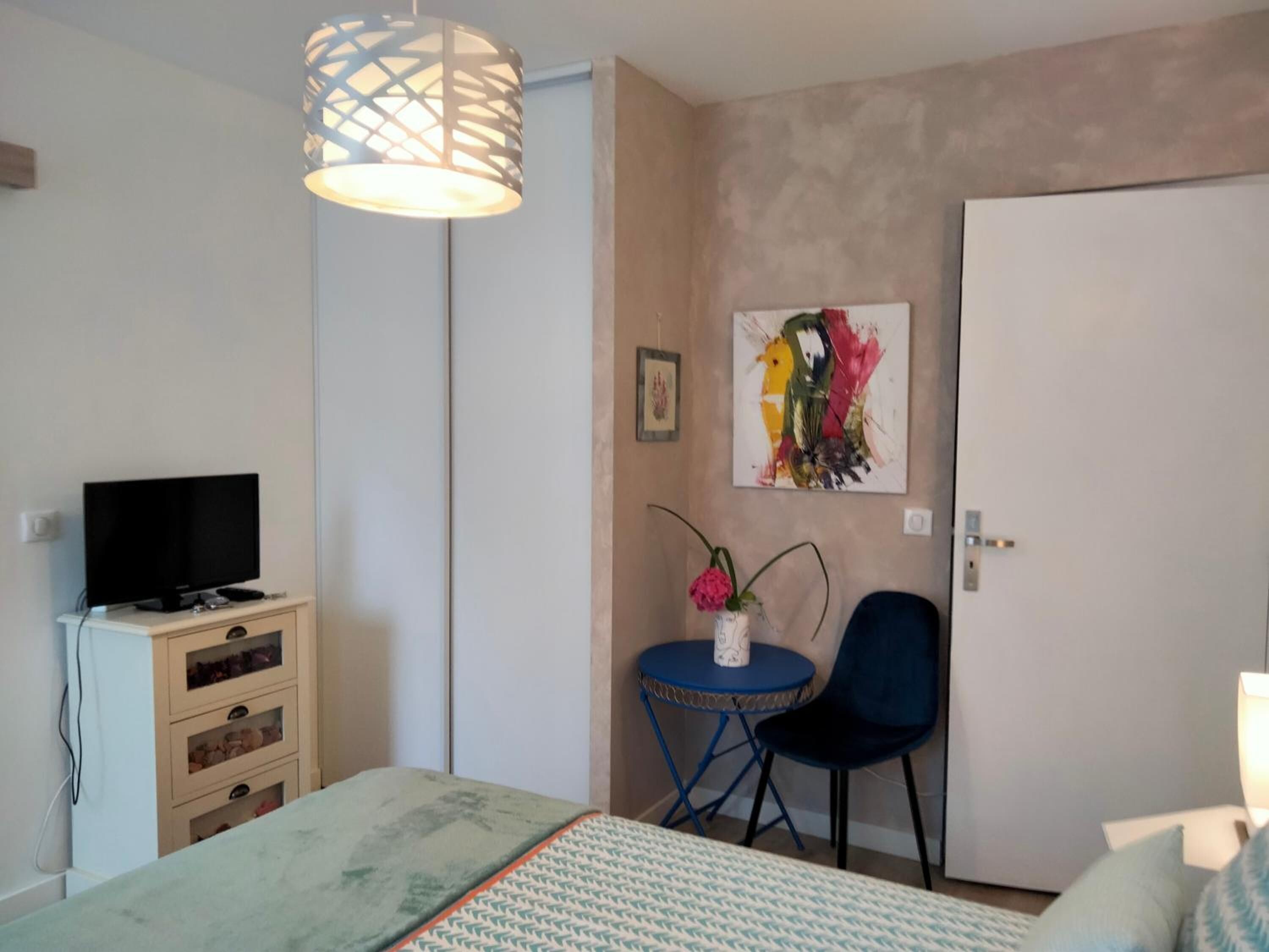 Un dormitorio con cama, televisor en una cómoda, escritorio con silla, cuadro en la pared y una puerta.