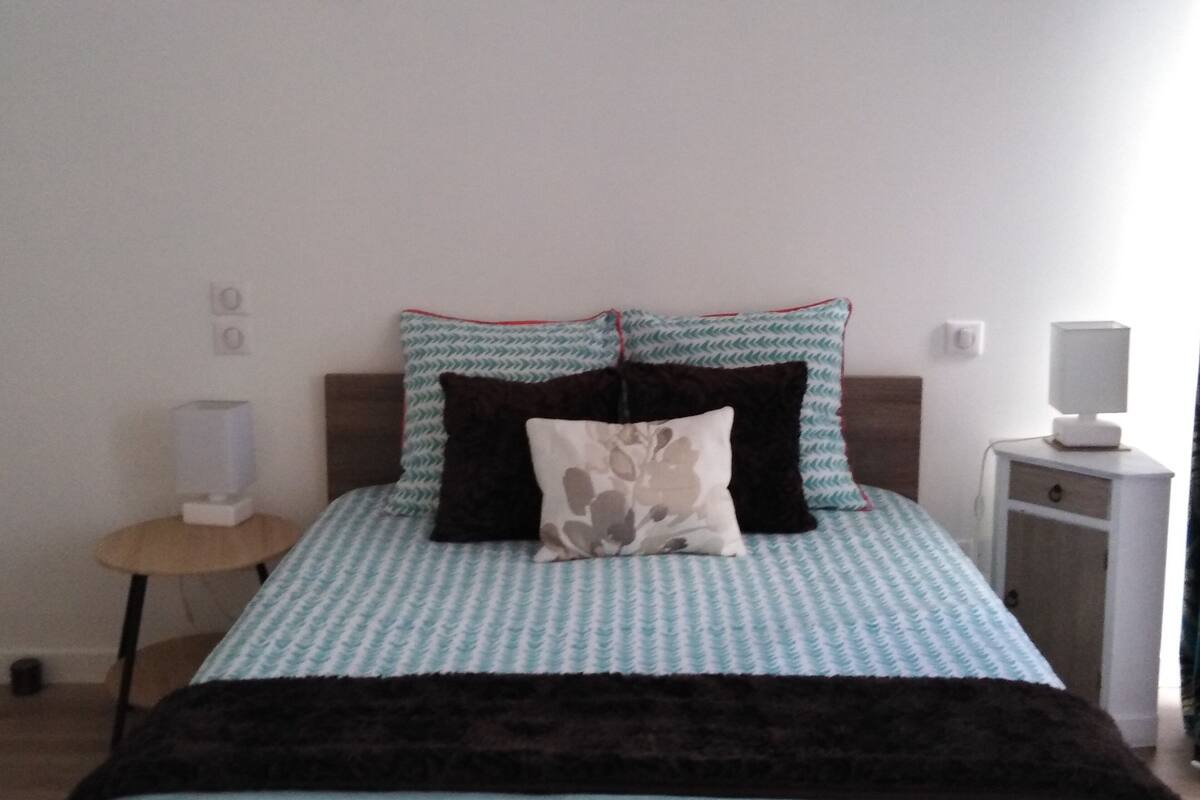 Una cama con una colcha estampada, dos almohadas, una mesita de noche con una lámpara y un buró con lámpara.