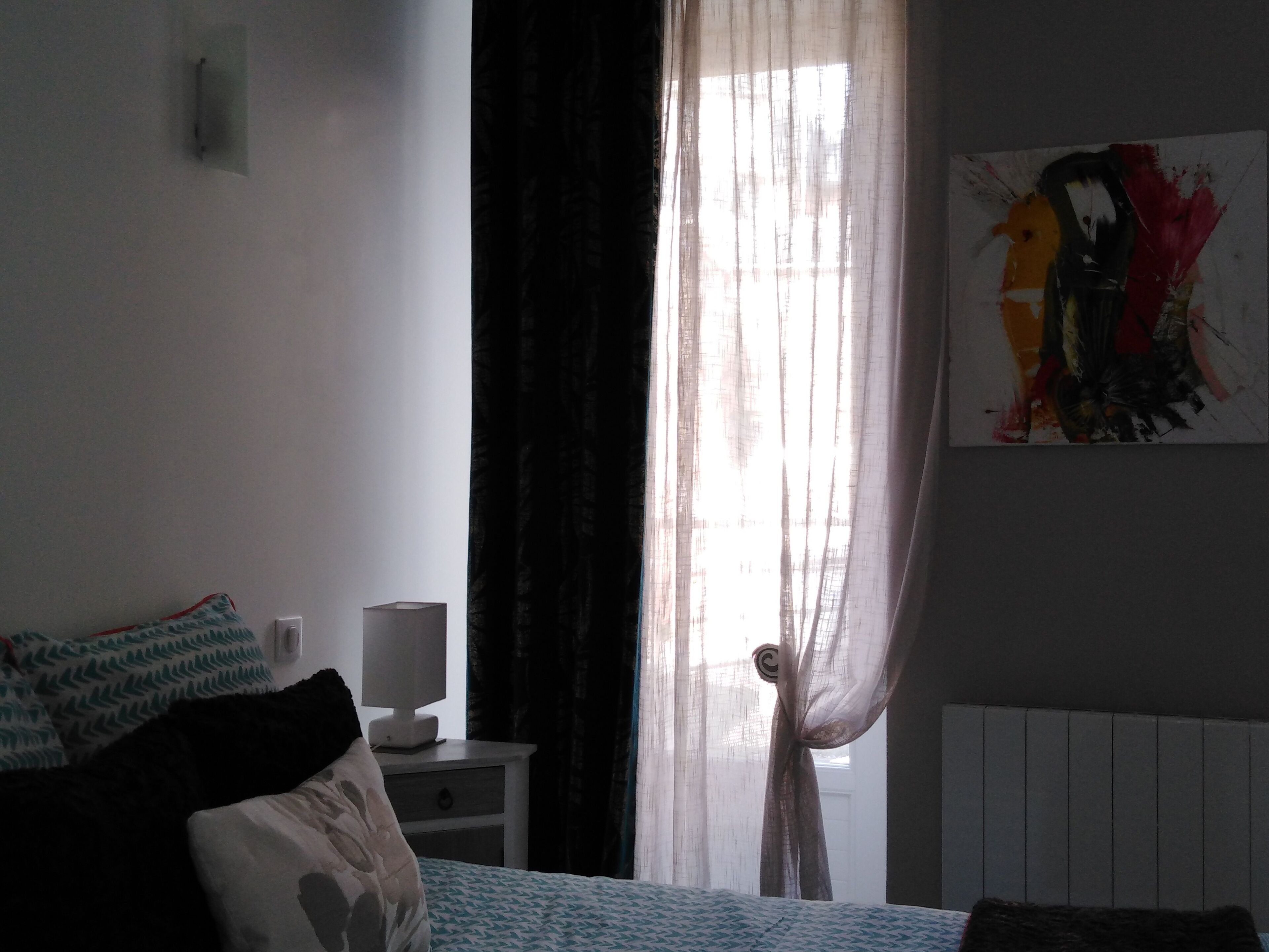 Una camera da letto con un letto, un comodino con una lampada, una finestra con tende leggere e un quadro appeso al muro.