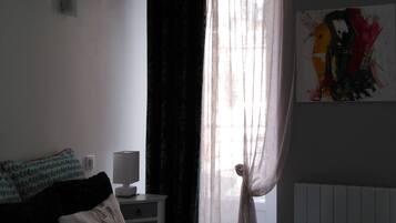 Un dormitorio con cama, una mesita de noche con lámpara, una ventana con cortinas translúcidas y un cuadro en la pared.