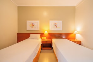 Quarto de hotel com duas camas de solteiro, cabeceiras de madeira, mesas de cabeceira com abajures e duas obras de arte emolduradas na parede.