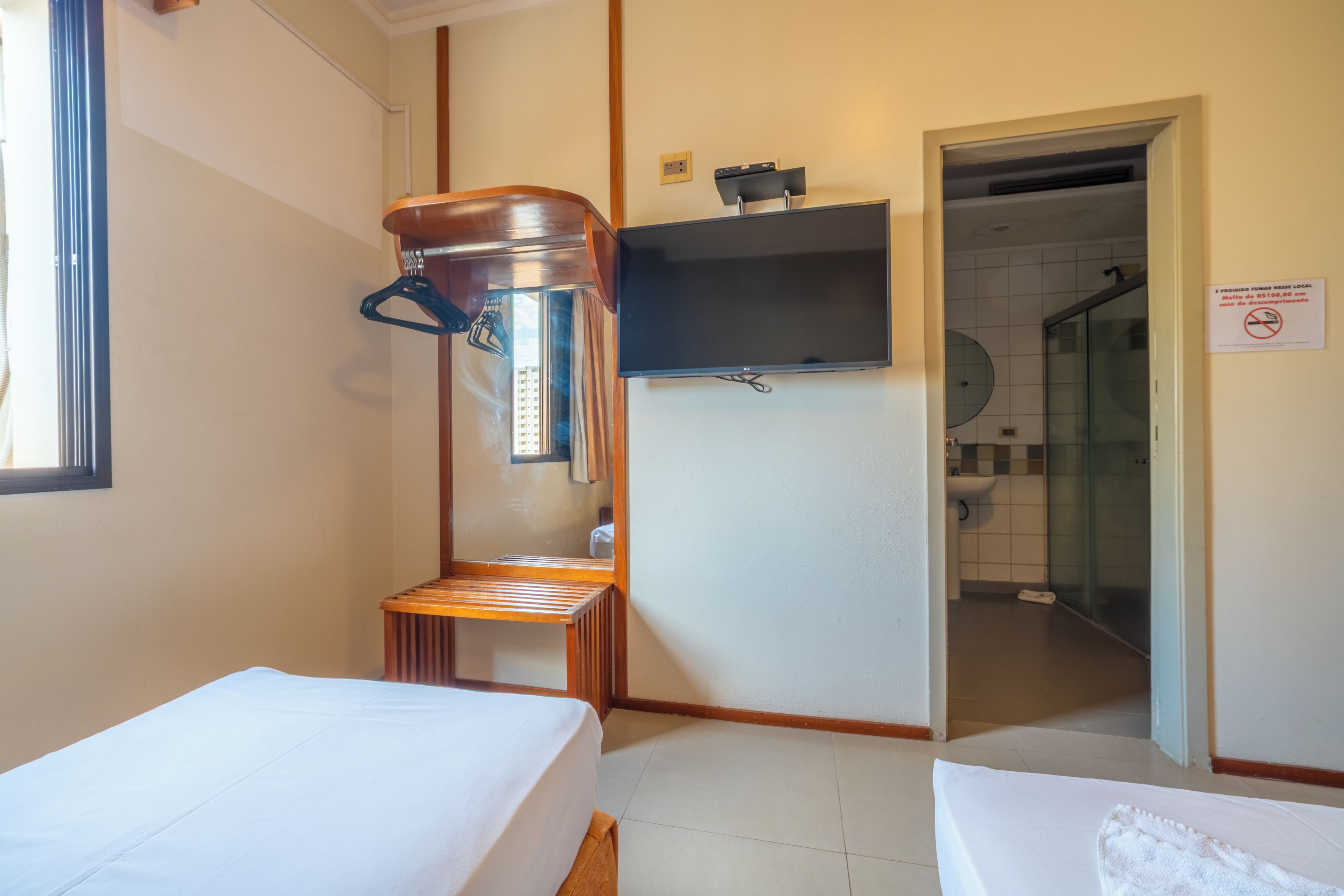 Una camera d'albergo con due letti, una sedia di legno, un televisore montato a parete e un box doccia visibile attraverso una porta aperta.