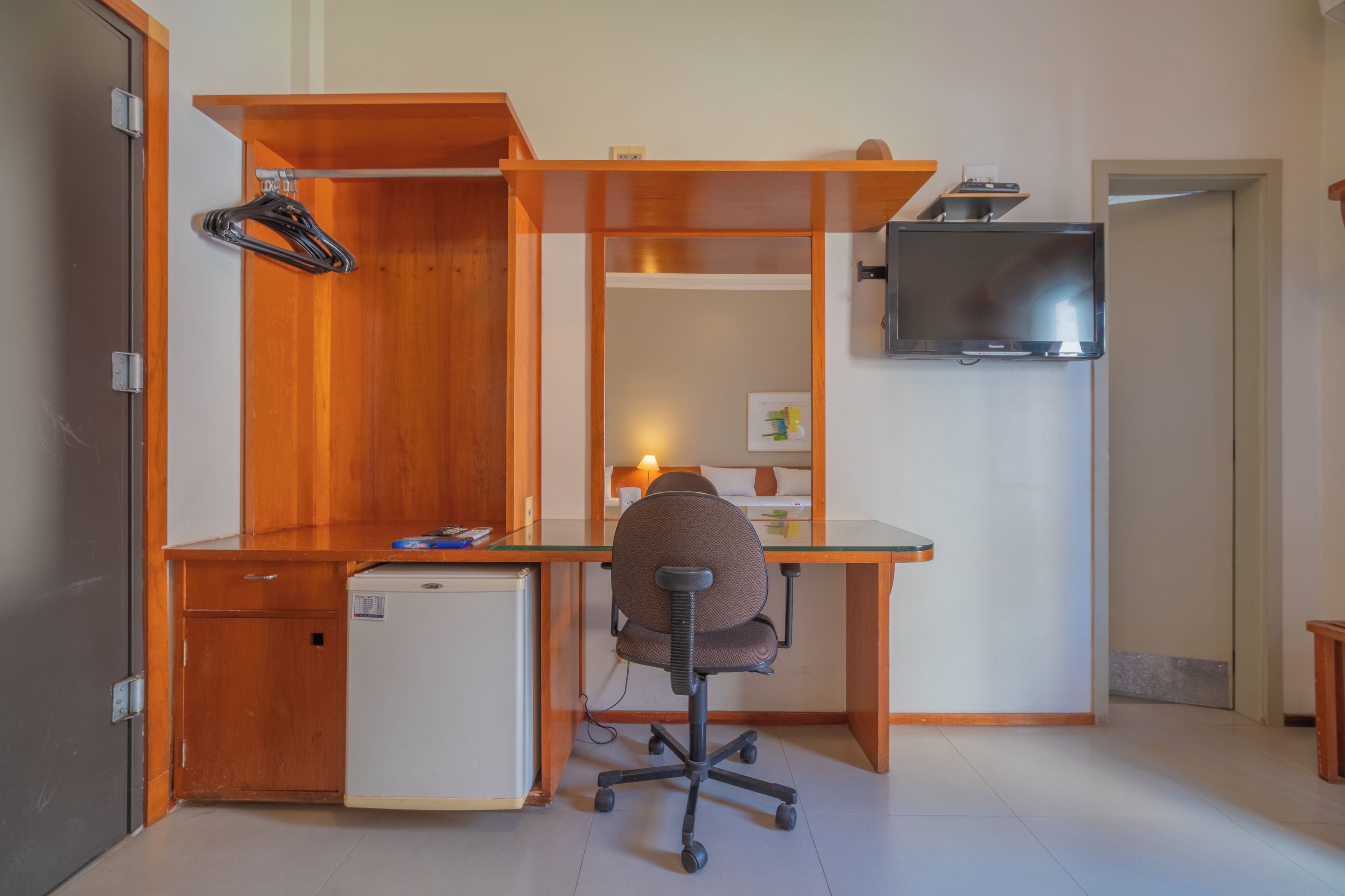A compact room with a wooden desk, a chair, a small refrigerator, a TV mounted on the wall, and a door leading to another room.