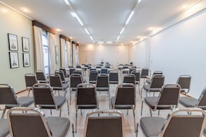 Sala de conferência com fileiras de cadeiras, uma tela de projetor e quadros nas paredes.