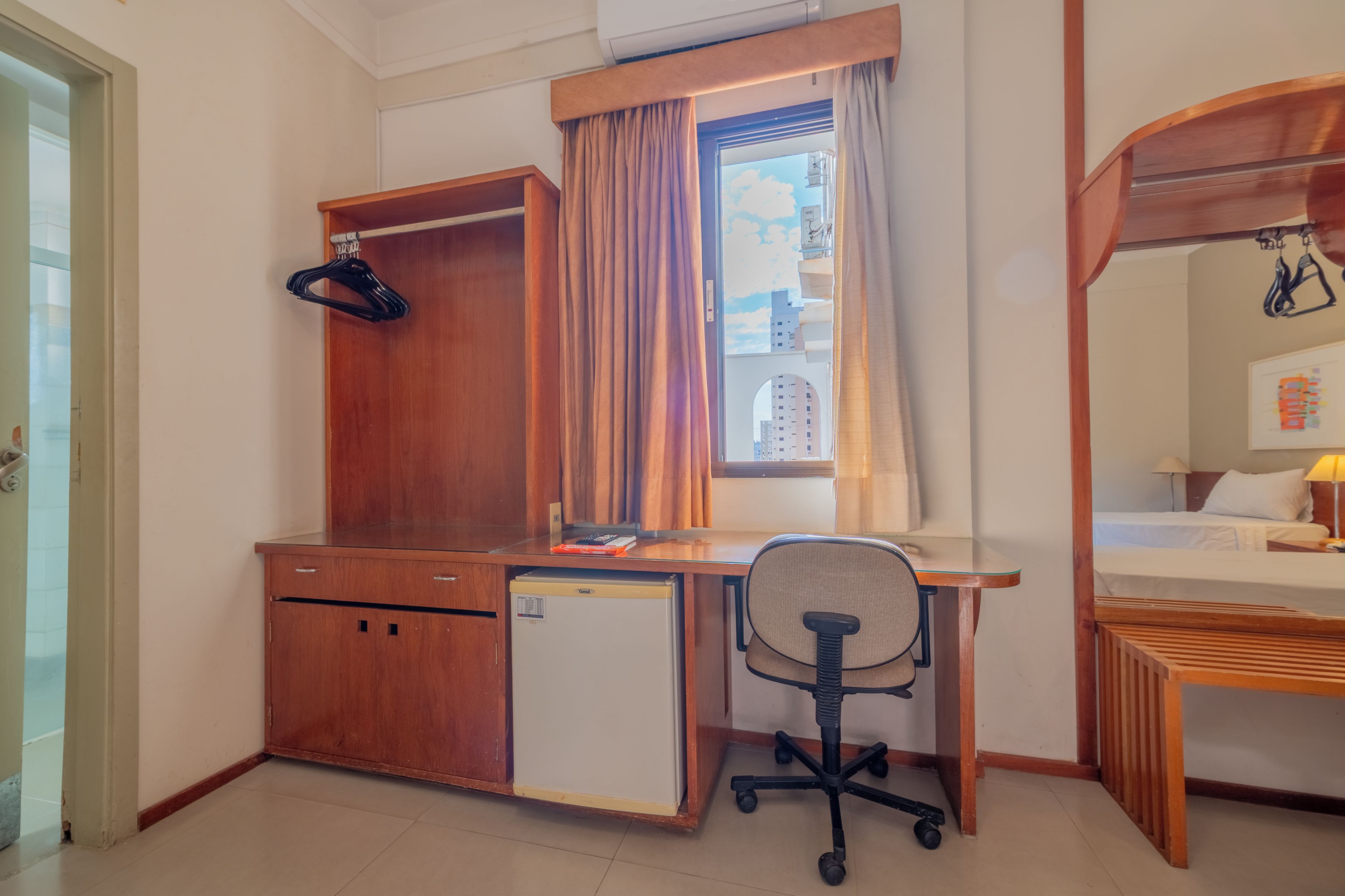 A compact hotel room with a wooden desk, a small refrigerator, a chair, a bed, and a window with curtains.