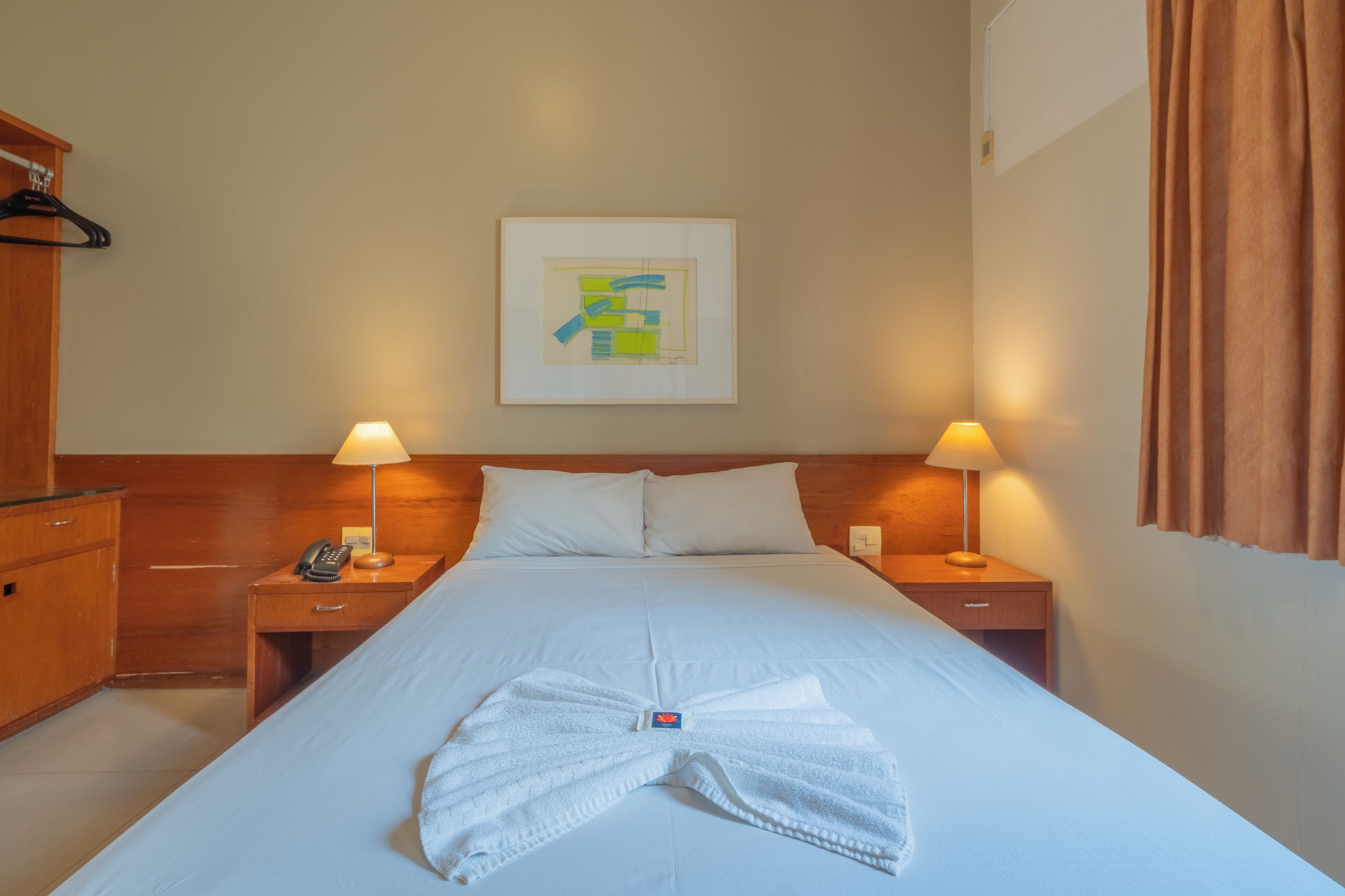 Une chambre d’hôtel avec un lit soigneusement fait, deux lampes de chevet, un téléphone, une table de chevet en bois et une œuvre d’art abstraite encadrée accrochée au mur.