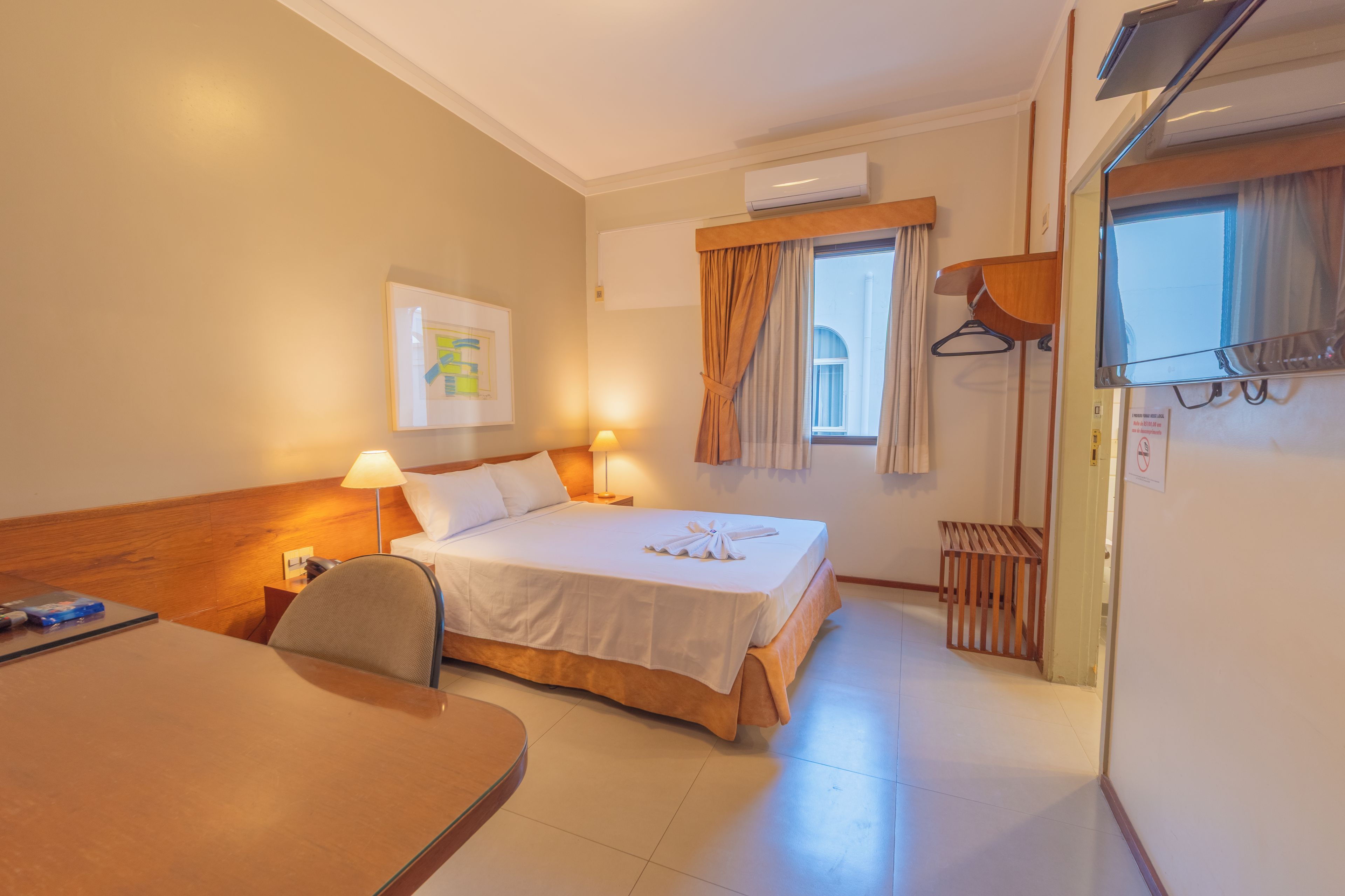 Een hotelkamer met een bed, nachtlampjes, een bureau, een stoel, een raam met gordijnen, een airconditioning en een wandkapstok.