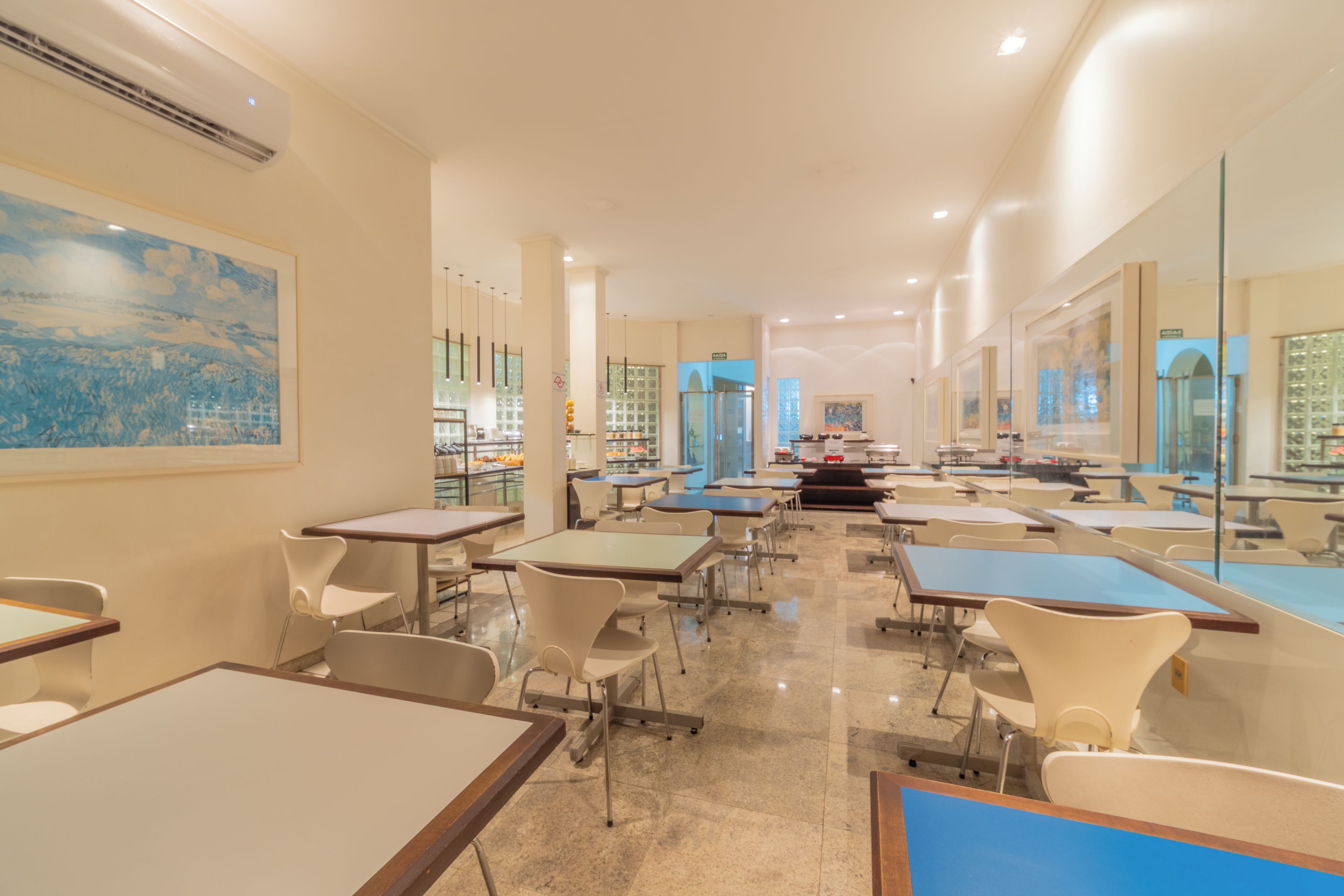 Un vaste espace de salle à manger avec plusieurs tables et chaises, un grand tableau au mur et une vue sur la cuisine.