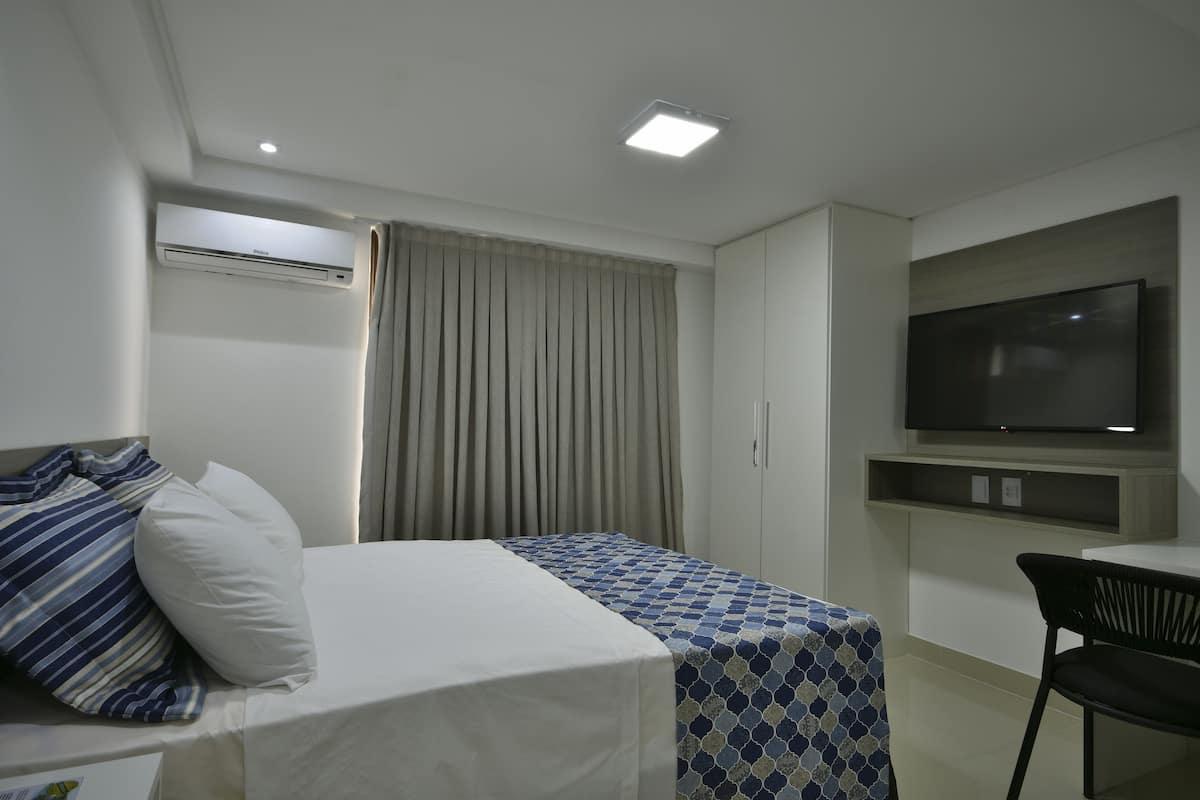 A hotel room with a bed, a TV mounted on the wall, and an air conditioning unit.