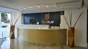 A reception desk with a logo, a flat-screen TV, and two decorative vases. - Littoral Gold Flat (João Pessoa)