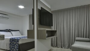 A modern hotel room with a bed, a sofa, a flat-screen TV mounted on the wall, and a large window with curtains. - Littoral Gold Flat (João Pessoa)