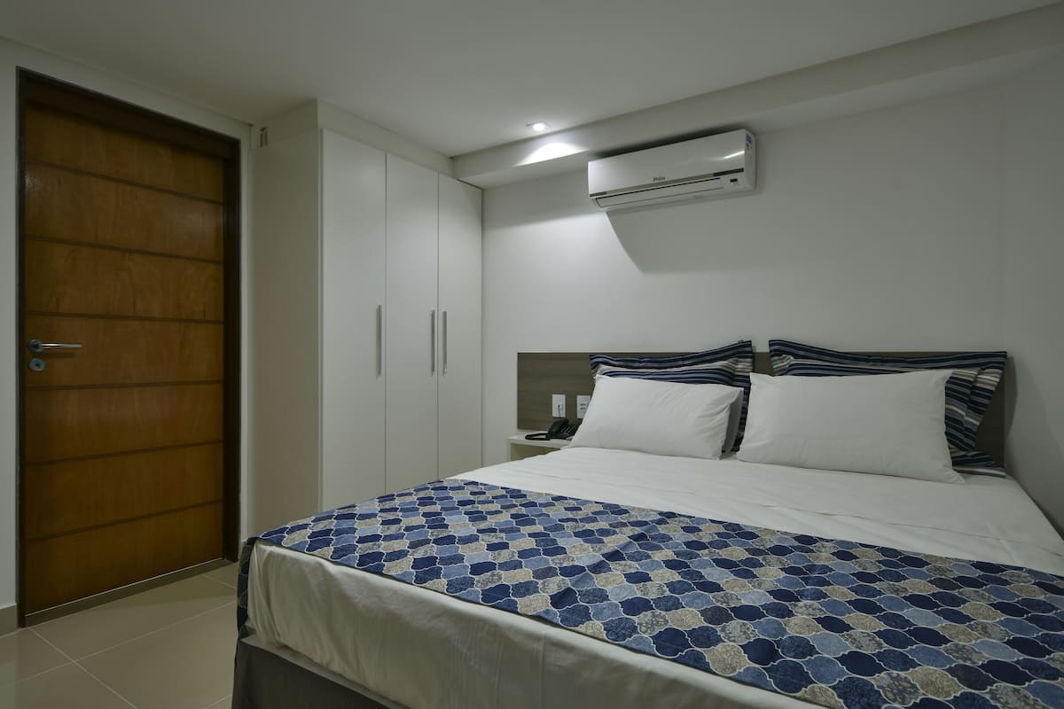 A bedroom with a bed, a wooden door, white cabinets, a nightstand, and an air conditioning unit.