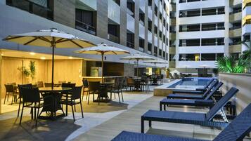 Un bar en la azotea con mesas al aire libre, piscina y un edificio moderno al fondo.
