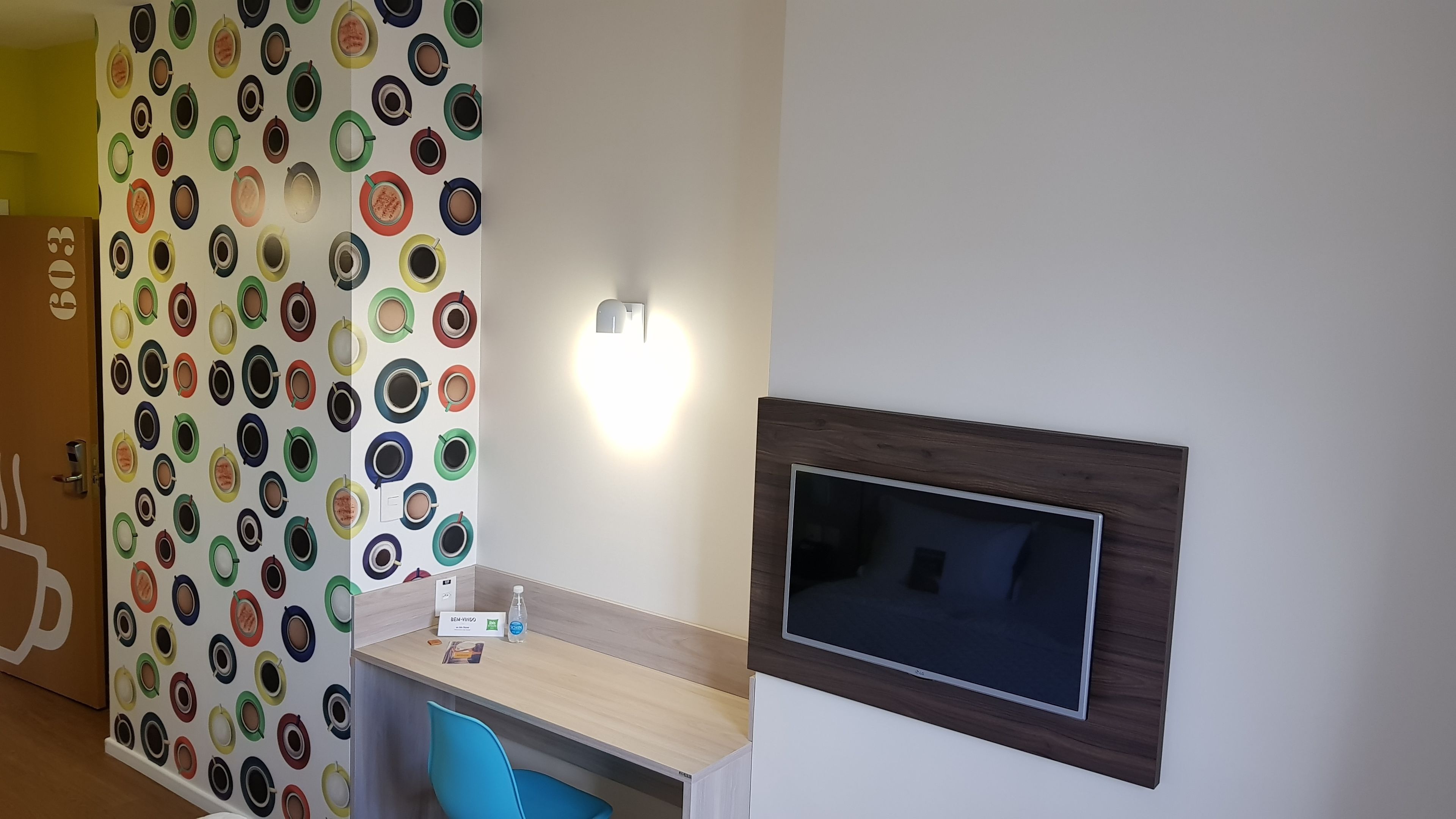 Una habitación con una pared decorada con tazas de café coloridas, un escritorio con una silla, un televisor de pantalla plana montado en la pared y una lámpara.
