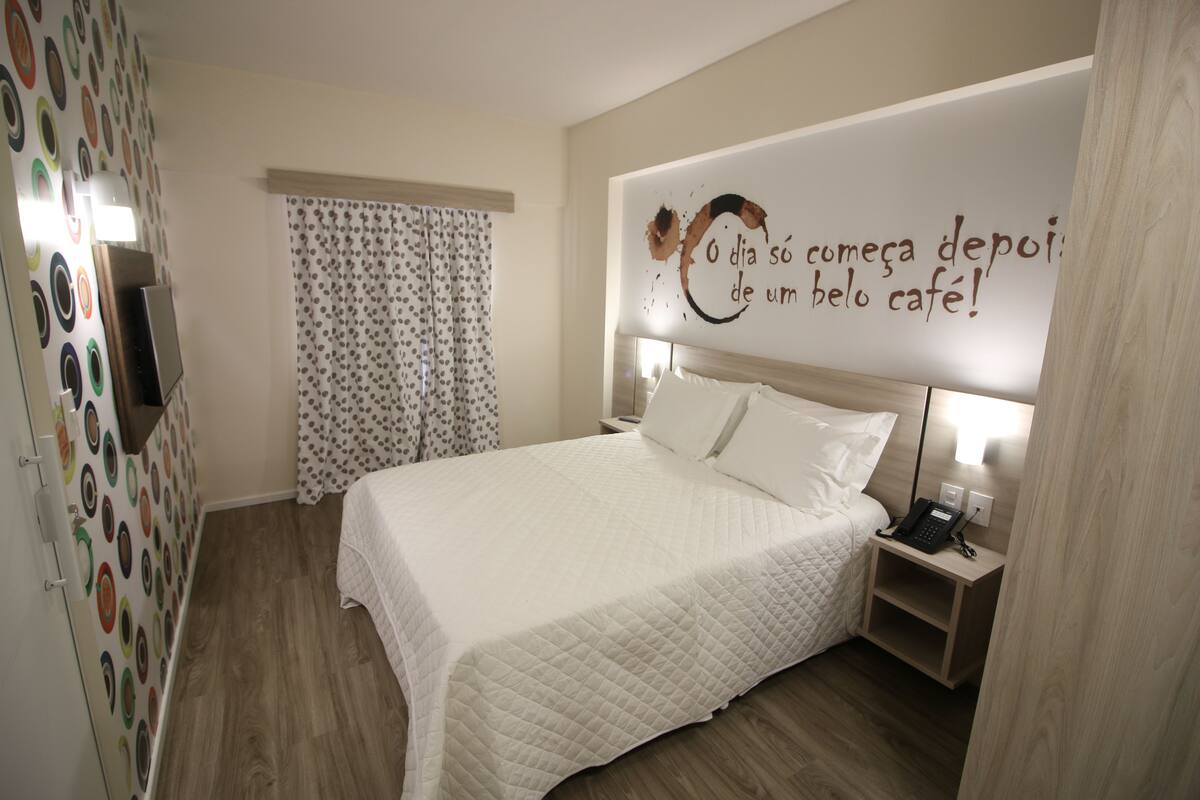 Una cama bien hecha con colcha blanca, una mesita de noche con un teléfono y una pared con una frase motivacional.