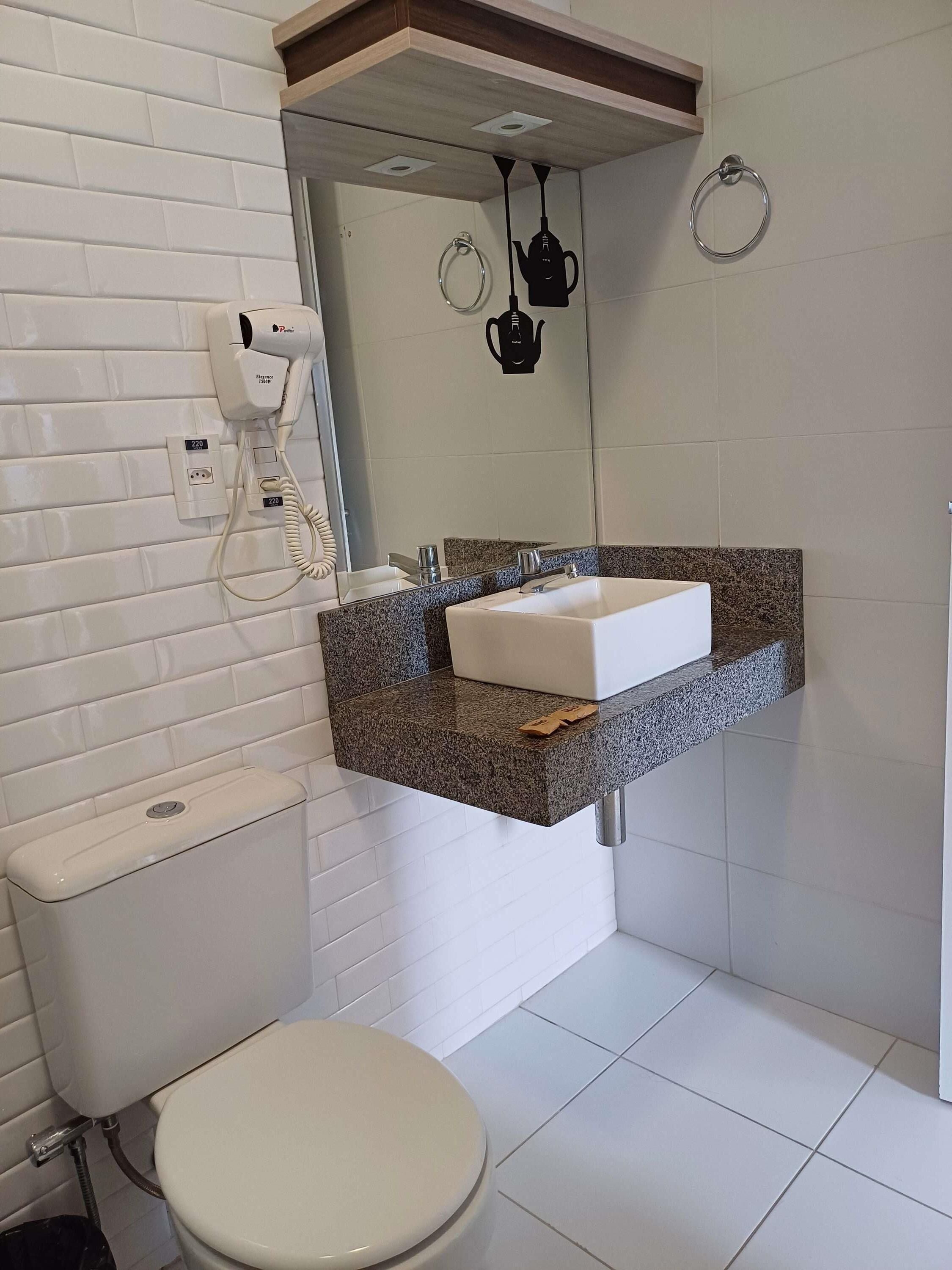 A bathroom with a white toilet, a granite countertop sink, a mirror, and a hairdryer mounted on the wall.