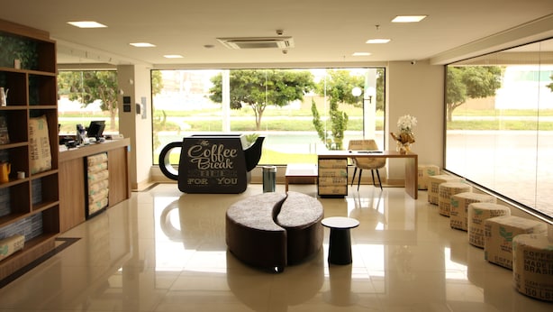 Un interior de cafetería moderna con un amplio ventanal, una estación de café, zona de estar y una estantería.