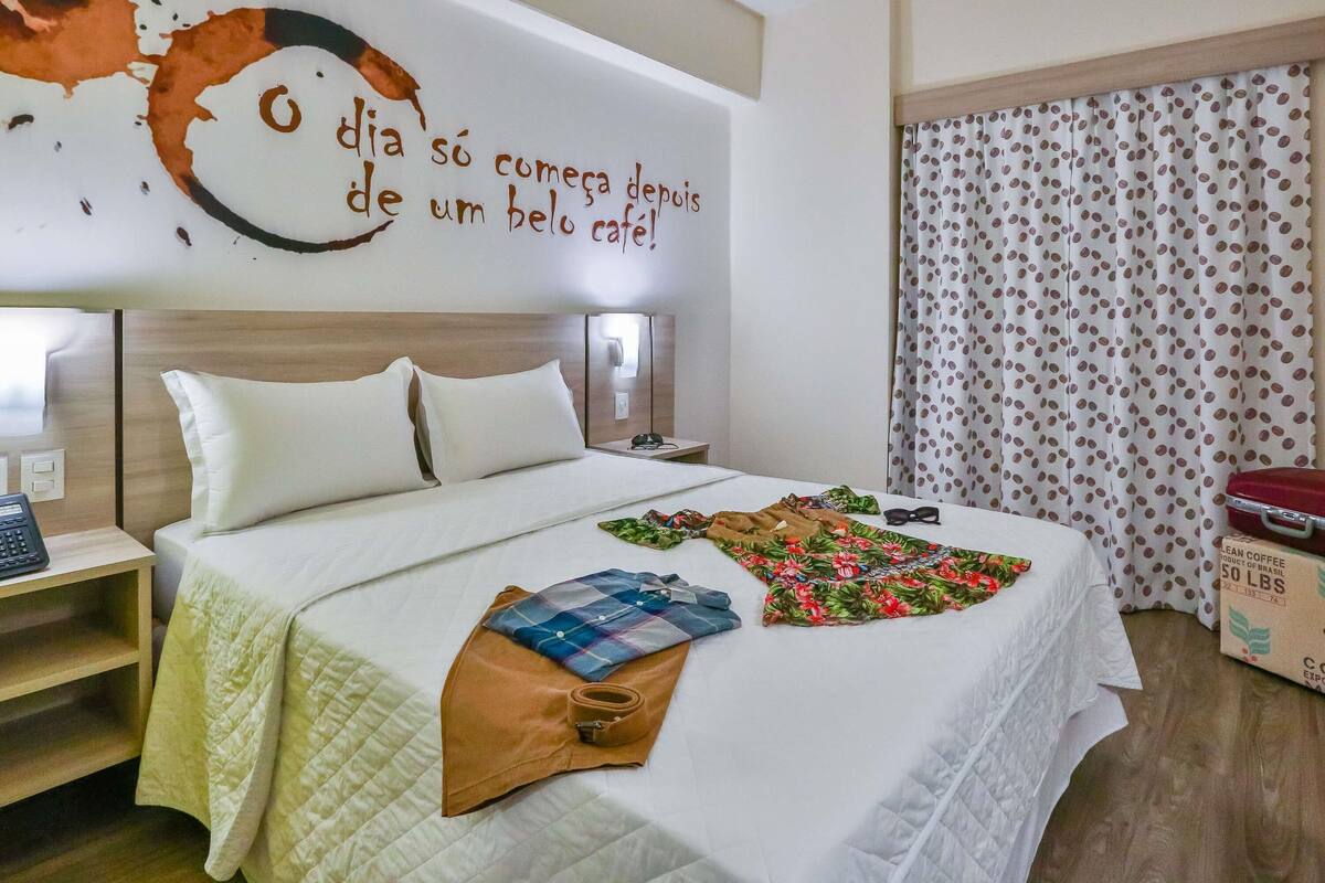 Una habitación de hotel con cama, mesita de noche, un cuadro con motivos de café, cortina y maleta.