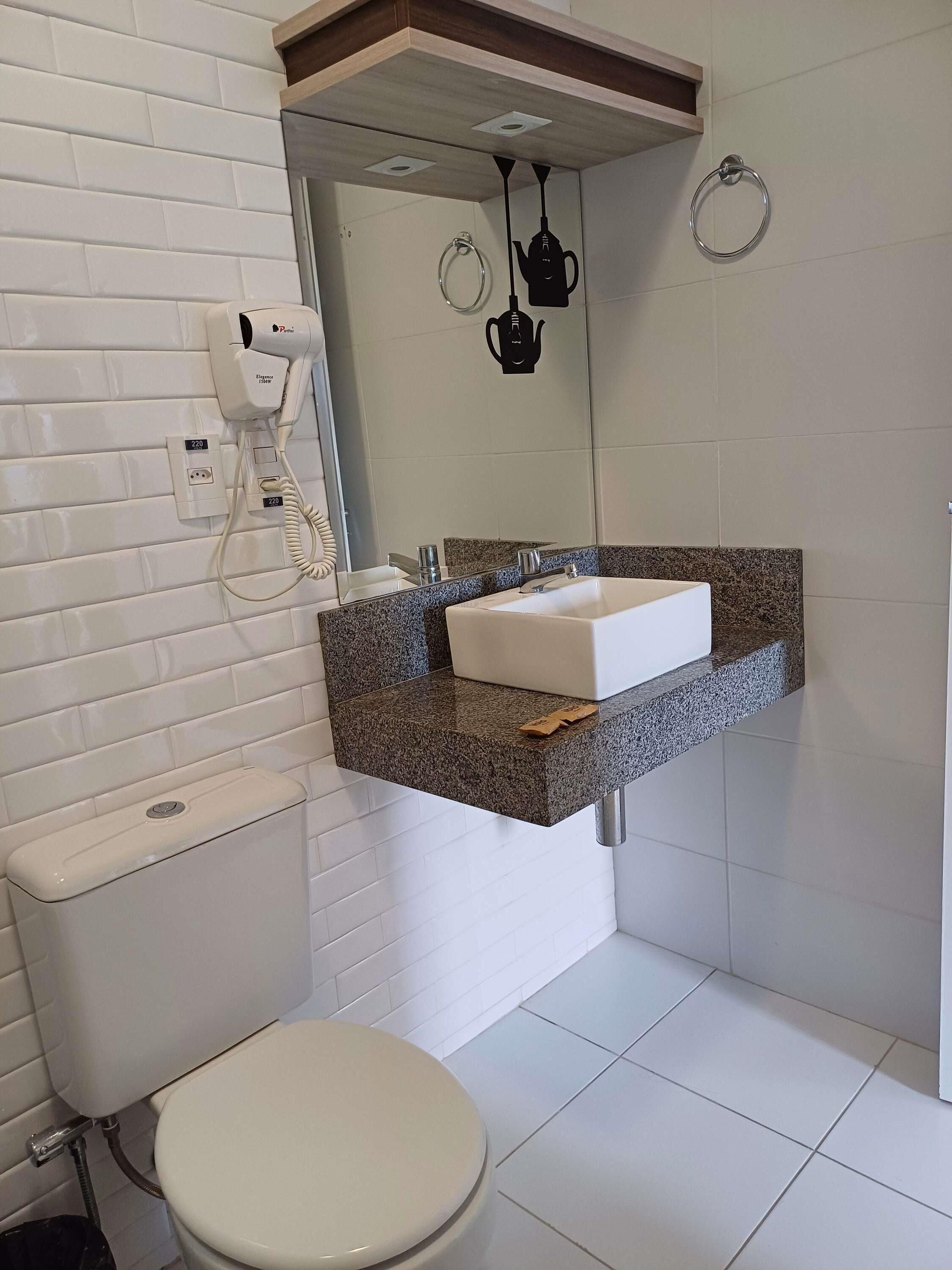 Un baño con inodoro blanco, lavamanos con encimera de granito, espejo y secador de pelo montado en la pared.