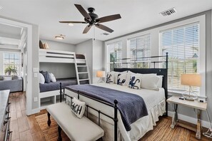 Une chambre avec un lit superposé, un bureau, une chaise, un banc, un ventilateur de plafond et de grandes fenêtres.