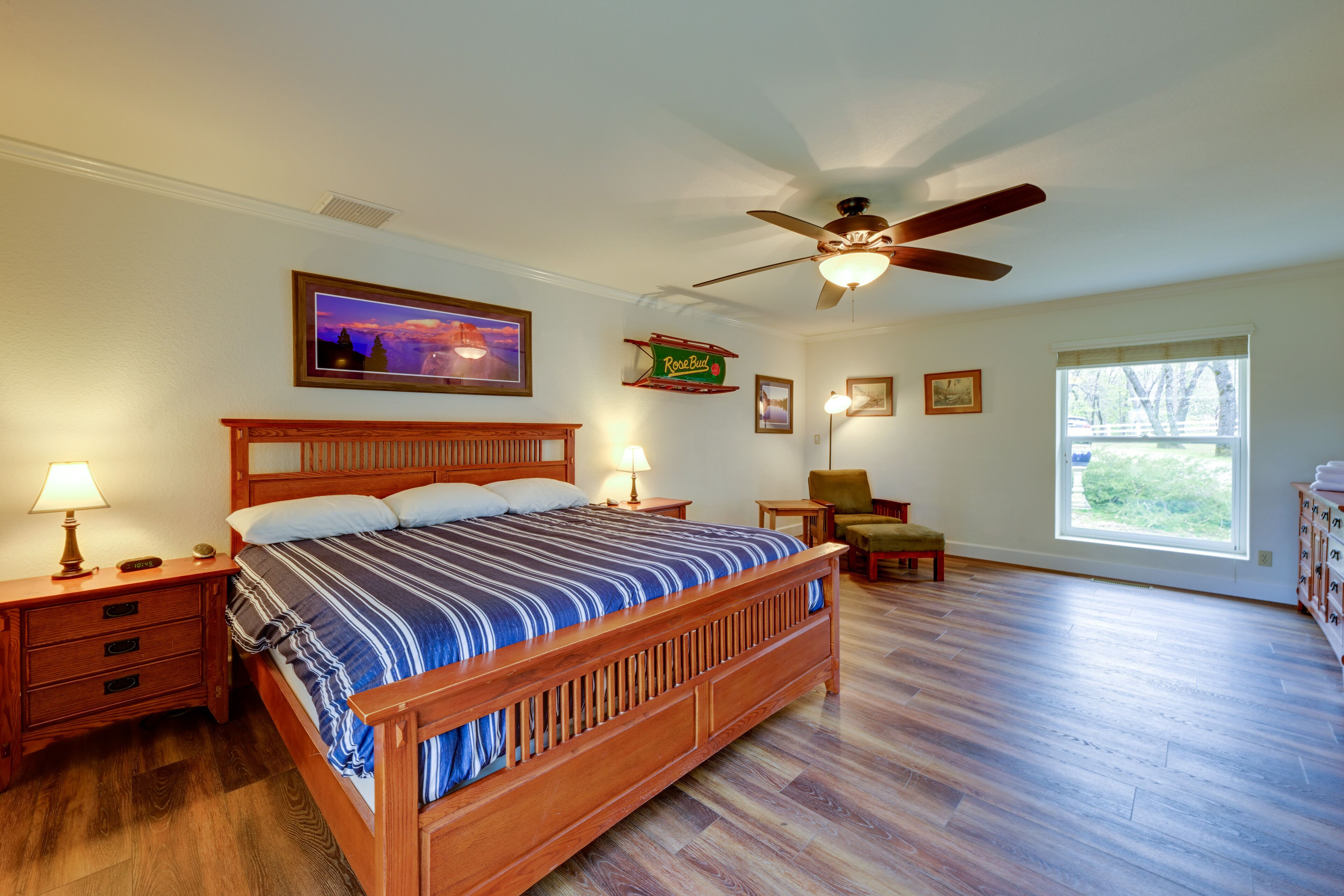 Quarto com cama de madeira, ventilador de teto, janela com vista e mesa de cabeceira com abajur.