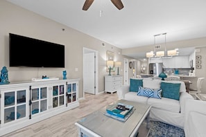 A modern living room with a flat-screen TV, a glass coffee table, and a white sofa with blue cushions.