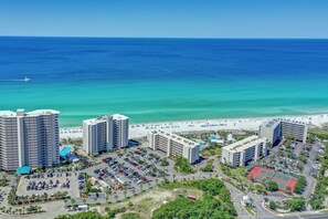 A coastal hotel with multiple high-rise buildings, a parking lot, and a beach in the background.