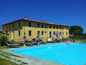 A pool area with lounge chairs and umbrellas, a yellow building with a tiled roof, and a clear blue sky. - Apartment in Palazzo- Residence Giuly Rosselmini -Pool (Pieve Santa Luce)