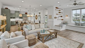 A spacious living room with a large window, a cozy seating area, and a kitchen in the background.