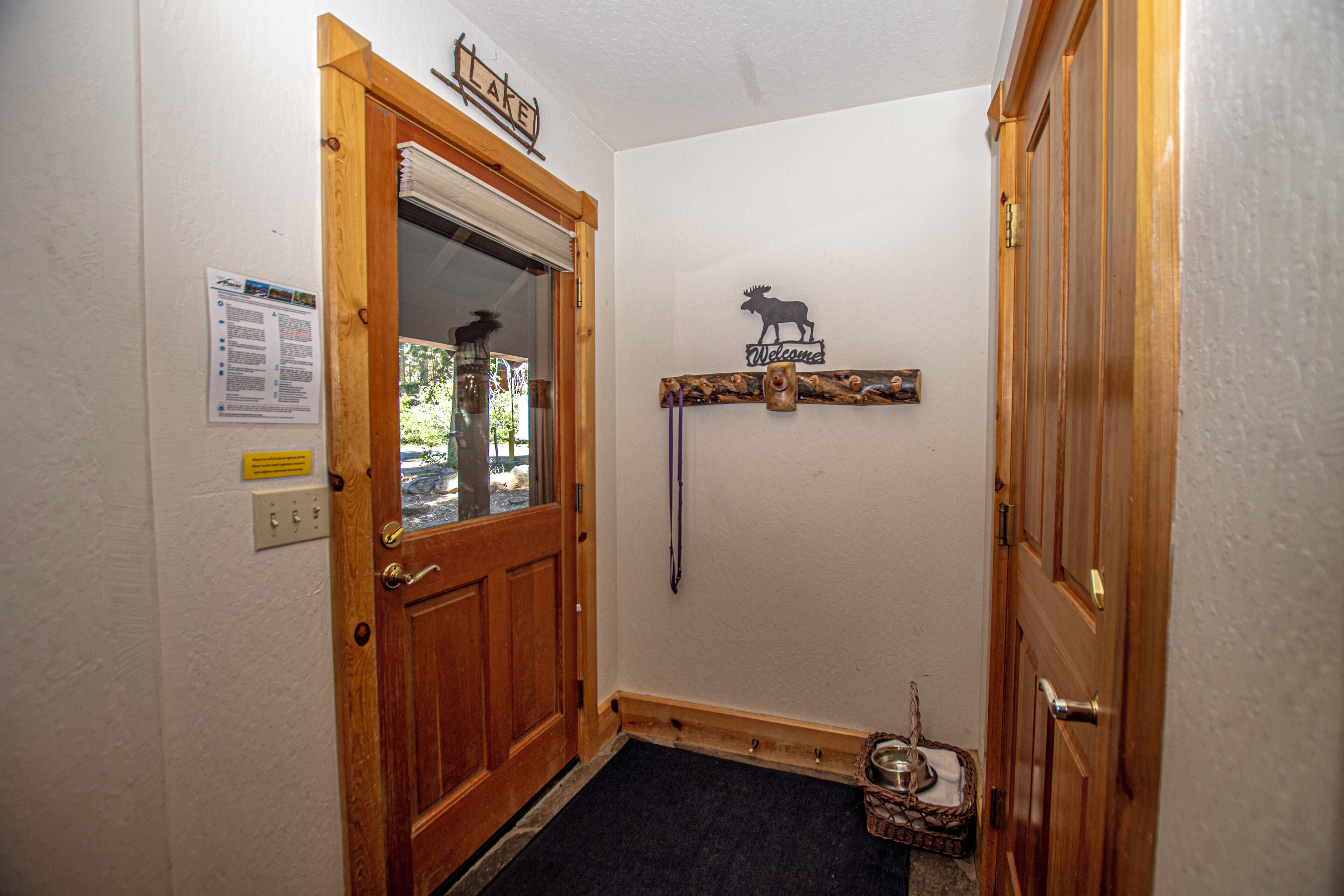 Una puerta de madera con un ventanal, un letrero de bienvenida con un alce, un estante de madera con un letrero y una pequeña cesta con un comedero para perro.