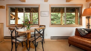 A dining area with a wooden table and chairs, a vase with flowers, and large windows with a view of trees. - RMR: La Choumine 6 Condo in Teton Village / Steps from the Mountain! (Teton Village)