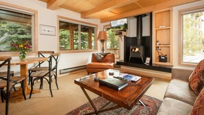 A cozy living room with a fireplace, wooden furniture, and large windows. - RMR: La Choumine 6 Condo in Teton Village / Steps from the Mountain! (Teton Village)