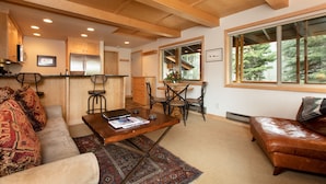 A cozy living room with a wooden ceiling, a dining area with a wooden table and chairs, and large windows offering a view of trees. - RMR: La Choumine 6 Condo in Teton Village / Steps from the Mountain! (Teton Village)