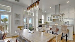 A modern kitchen with white cabinets, stainless steel appliances, and a dining area with a wooden table and wicker chairs. - Beautiful Beach Home in Cinnamon Shore w/Gulf Peeks & Access to Amenities (Port Aransas)