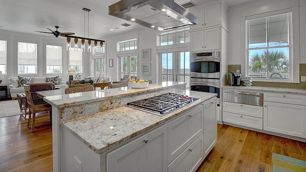 A modern kitchen with white cabinetry, a large island with a granite countertop, stainless steel appliances, and a view of palm trees through the window. - Beautiful Beach Home in Cinnamon Shore w/Gulf Peeks & Access to Amenities (Port Aransas)