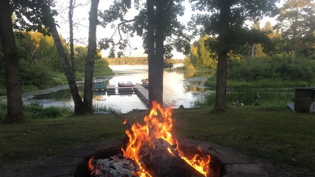 A campfire in a stone fire pit surrounded by grass and trees, with a body of water and dock in the background. - Luxury RV at Bolton Bay on Long Lake (Park Rapids)