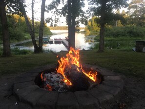 A campfire in a stone fire pit surrounded by grass and trees, with a body of water and dock in the background.