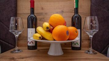 Una mesa de madera con un soporte blanco para pasteles que sostiene naranjas y plátanos. Dos botellas de vino y dos copas de vino vacías están colocadas a ambos lados del soporte.