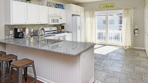 A kitchen with white cabinets, a granite countertop, a gas stove, and a sink. There are three bar stools and a sign that says "BEACH". - Water Street Townhouses (Saugatuck)