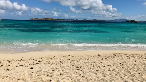 A beach with clear turquoise water, sandy shore, and distant islands under a partly cloudy sky.