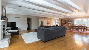 Een ruime woonkamer met een grijze modulaire bank, houten vloeren, een witte vloerkleed en een eethoek met houten stoelen en een tafel.
