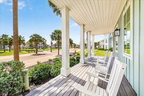 A covered porch with white wooden benches, palm trees, and a view of a street with buildings in the background. - Family-Size Beach Home in Cinnamon Shore w/ Access to Great Amenities (Port Aransas)