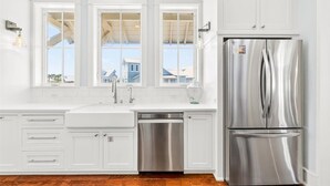 Une cuisine avec des meubles blancs, un réfrigérateur en acier inoxydable, un lave-vaisselle et une fenêtre donnant sur des maisons.