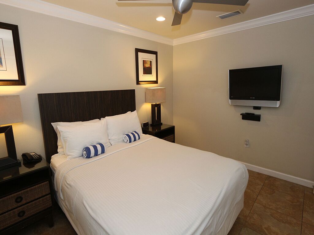Une chambre d’hôtel avec un grand lit, un téléviseur à écran plat, un ventilateur de plafond et un tableau encadré au mur.