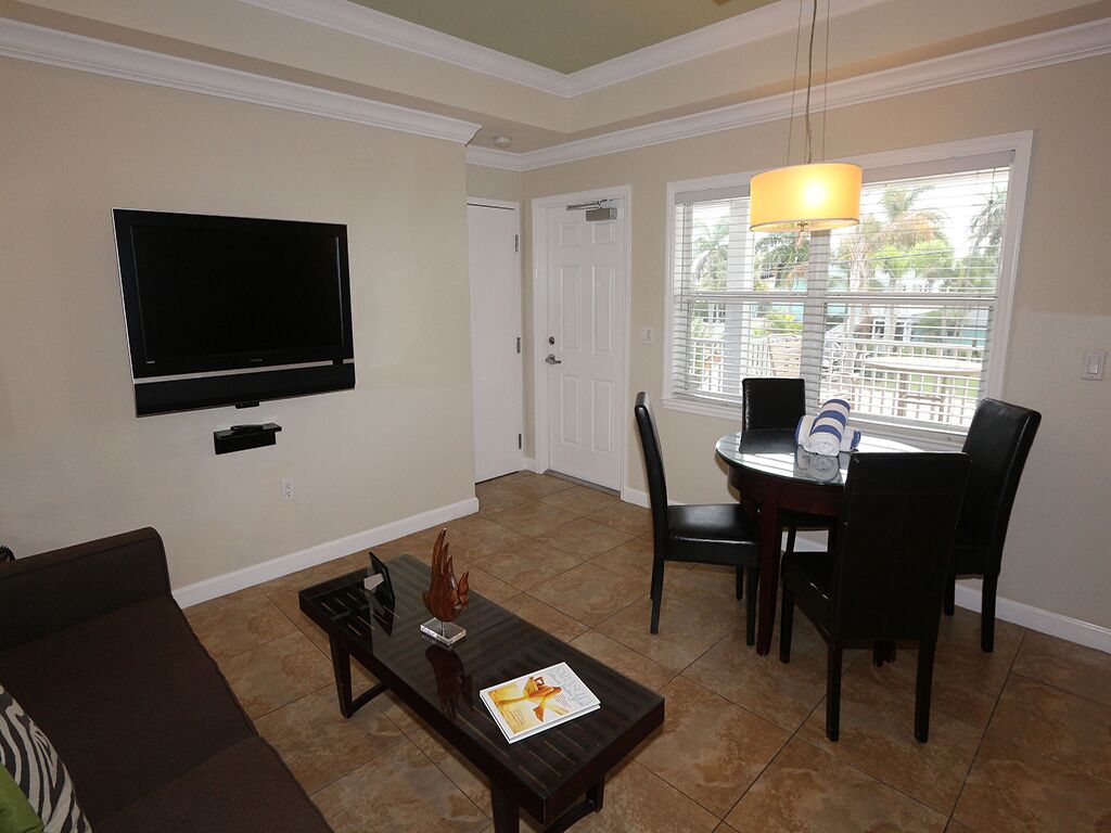 A living room with a flat-screen TV mounted on the wall, a dining table with chairs, a coffee table with a magazine, and a window with blinds.
