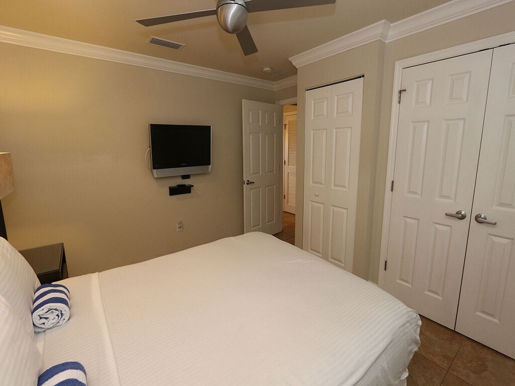 A hotel room with a bed, a ceiling fan, a TV mounted on the wall, and white doors.